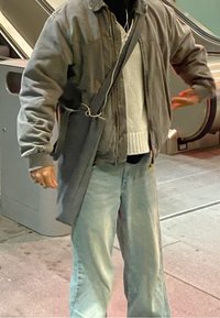 Giacca over-size grigia sopra un maglione chiaro, abbinata a jeans larghi di un blu chiaro. Una borsa a tracolla grigia con una cinghia poggia contro il corpo.