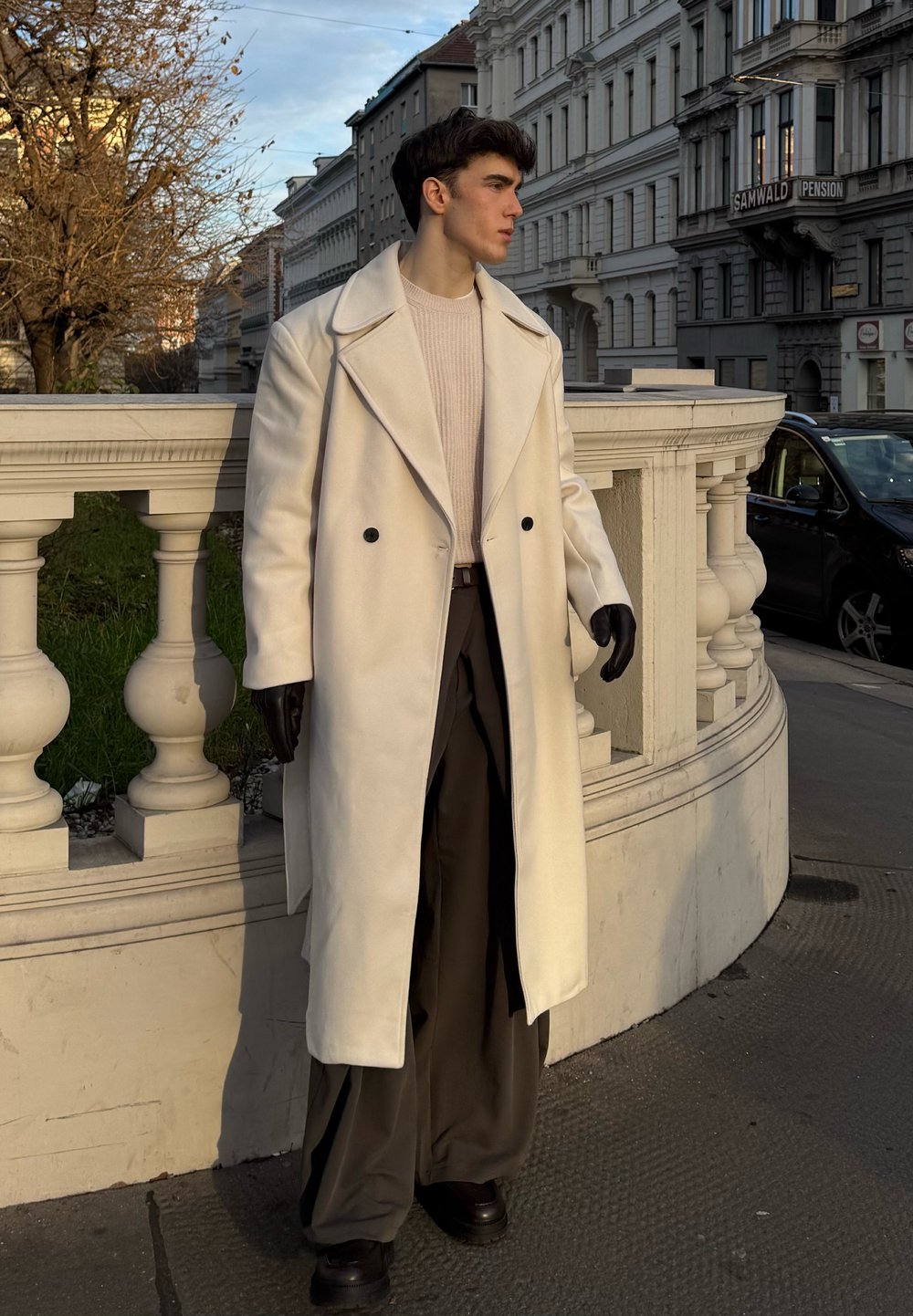 Long manteau blanc avec grands revers, boutons noirs, porté avec un pull léger et un pantalon large foncé. Gants noirs et chaussures épaisses. Cadre urbain.