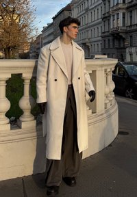 Long manteau blanc avec grands revers, boutons noirs, porté avec un pull léger et un pantalon large foncé. Gants noirs et chaussures épaisses. Cadre urbain.