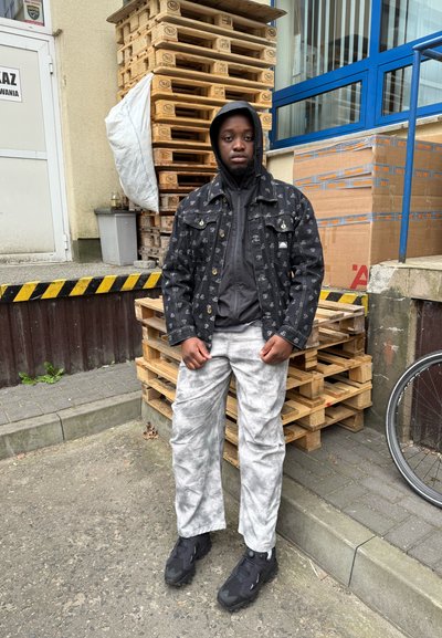 Joven con chaqueta negra con estampado y capucha, pantalones grises y zapatillas negras, de pie cerca de palets de madera apilados y cajas de cartón al aire libre.