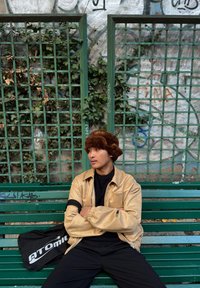 Personne assise sur un banc vert, portant une veste en similicuir beige et un pull noir, avec un chapeau en fausse fourrure marron et un sac noir affichant "ATOMIC".