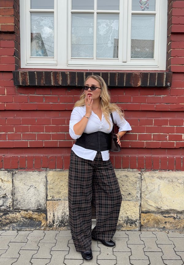 Femme aux longs cheveux blonds portant des lunettes de soleil noires, une chemise blanche, un corset noir, un pantalon large à carreaux et des chaussures noires, debout contre un mur de briques rouges.