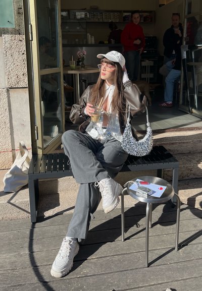 Joven mujer con gorra blanca y chaqueta marrón está sentada en un banco fuera de una cafetería, sosteniendo un café con hielo, con un bolso plateado y una mesa pequeña cerca.