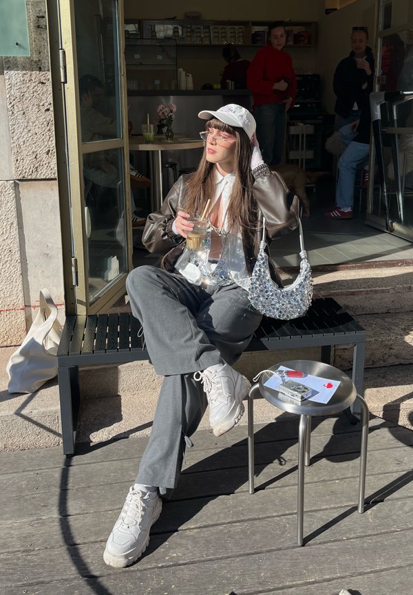 Mlada žena u bijeloj kapici i smeđoj jakni sjedi na klupi ispred kafića, drži ledenu kavu, sa srebrnom torbicom i malim stolom u blizini.
