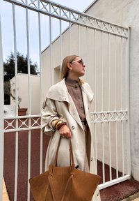 Cămașă bej peste un pulover maro, accesorizată cu cercei și brățări din aur. Ține o geantă din suede maro lângă o poartă albă.