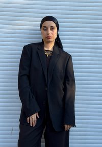 Črni oversized blazer z enim gumbom, nošen čez črno majico. Kombiniran z zlato verižico in usklajenim ruto za glavo. Teksturirana podlaga.