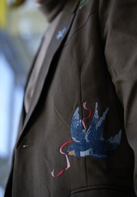 Blazer marrón con un intricado bordado de pájaro azul en la manga izquierda, con detalles de cintas rosas y rojas. Textura de tela suave.