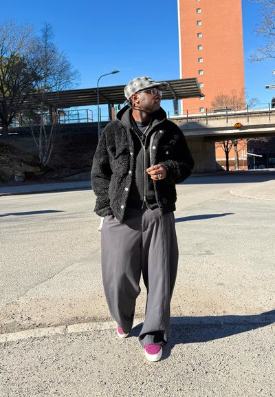 Hombre vestido con una chaqueta negra peluda, pantalones anchos grises, zapatillas con estampado, gorra gris y gafas caminando al aire libre cerca de un paso elevado en un día soleado.