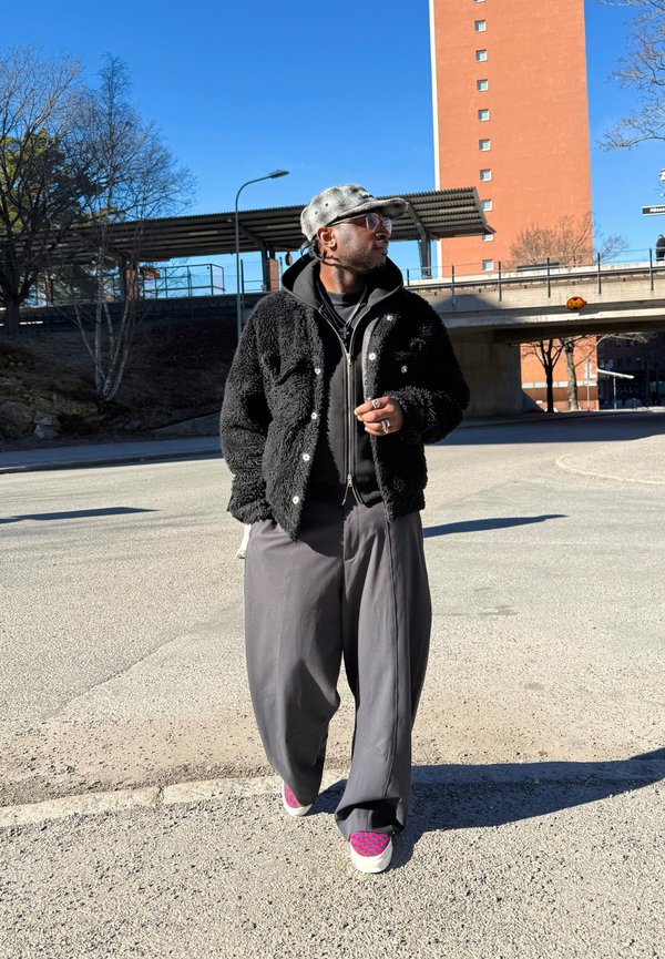 Homme portant une veste noire pelucheuse, un pantalon large gris, des baskets à motifs, une casquette grise et des lunettes, marchant à l'extérieur près d'un passage supérieur par une journée ensoleillée.