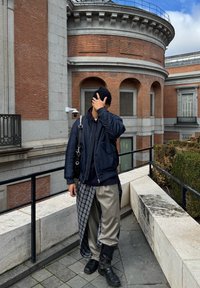 Ναυτική σακάκι, μαύρο σκουφί, καρώ πουκάμισο πάνω από κρεμ παντελόνι και μαύρες μπ�ότες μέχρι το γόνατο; στέκεται σε μια πέτρινη καγκελό.