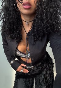 Cămașă neagră cu butoni, suprapusă peste un bralette din dantelă, decorată cu un colier multicolor și brățări metalice, asortată cu pantaloni negri cu franjuri.