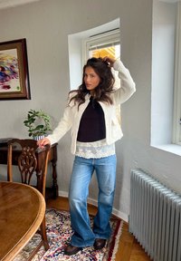 Mujer con cabello largo y oscuro, chaqueta blanca, camiseta negra y jeans azules, de pie junto a una silla de madera cerca de una ventana en una habitación acogedora.