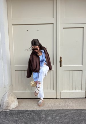 Mujer con gafas de sol se ajusta el zapato mientras está de pie frente a unas puertas beige con paneles, sosteniendo un pequeño bolso crema.