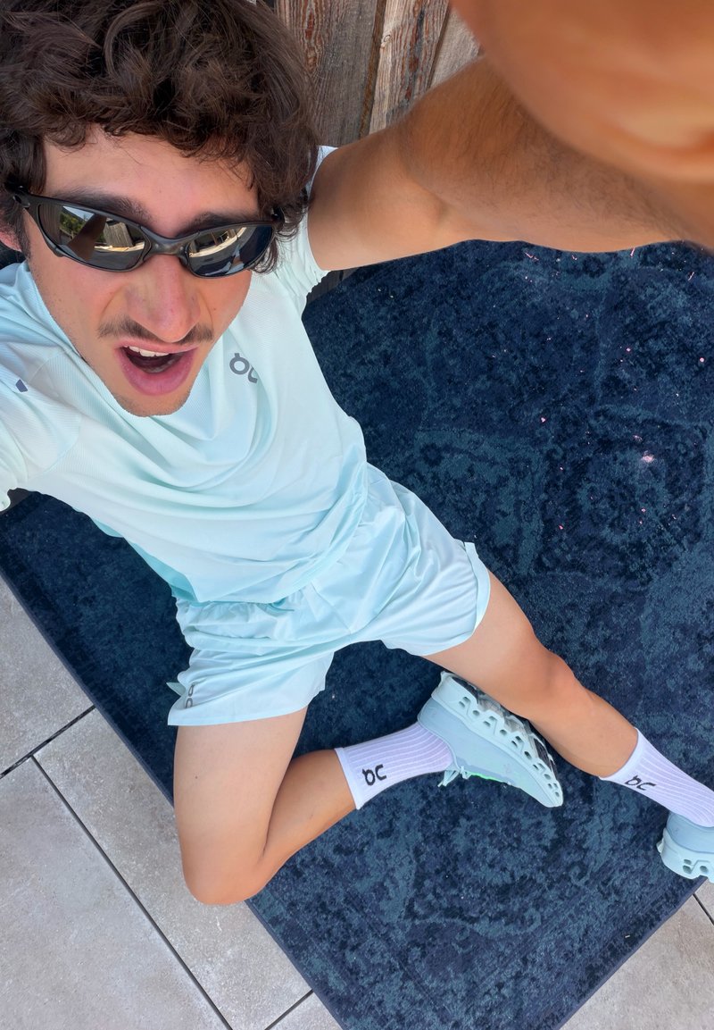 Jeune homme en tenue de sport bleu clair et lunettes de soleil assis sur un tapis bleu foncé sur un sol carrelé, prenant un selfie de dessus.