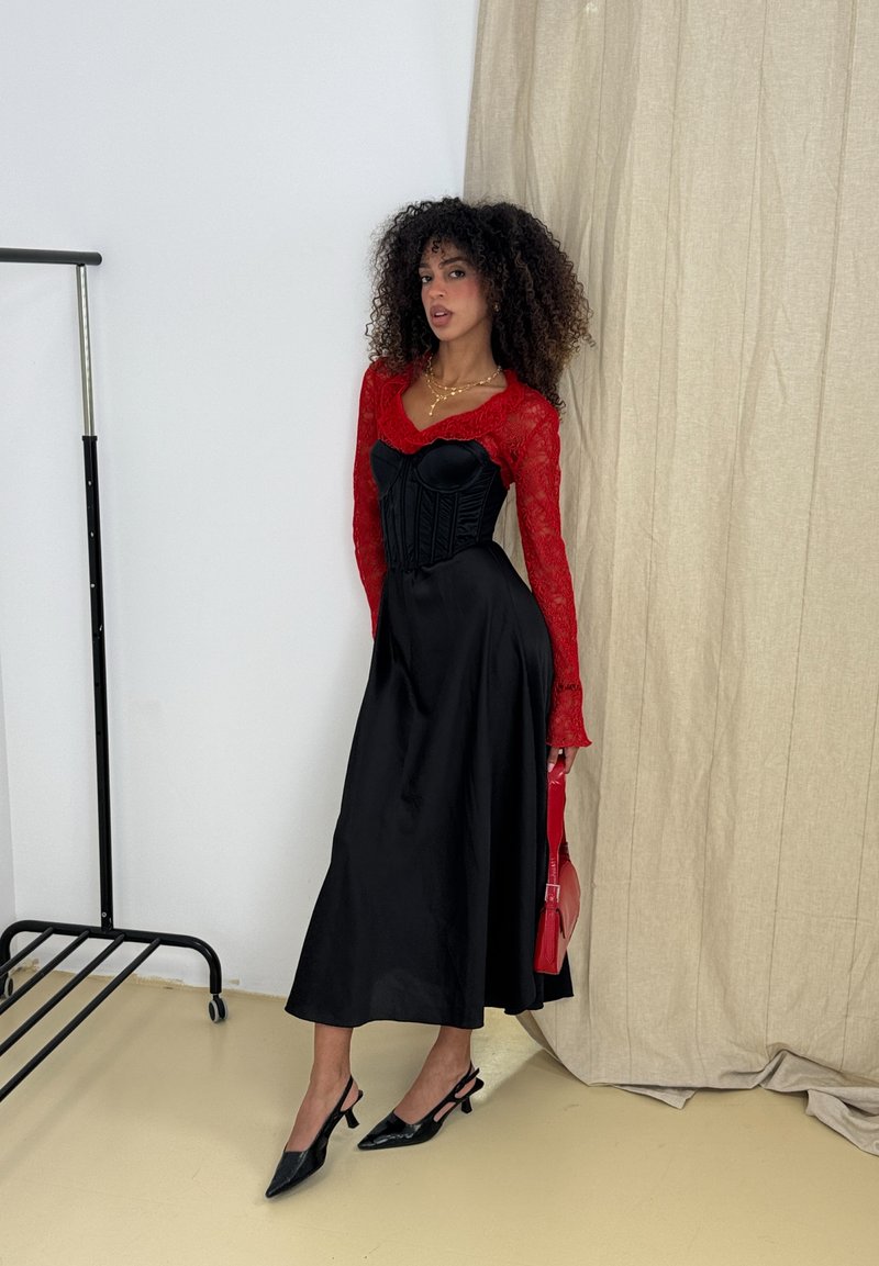 Vestido de cetim preto com um corpete justo e saia fluida, combinado com uma blusa de renda vermelha de mangas longas. Os acessórios incluem uma bolsa vermelha e sapatos de salto aberto pretos.