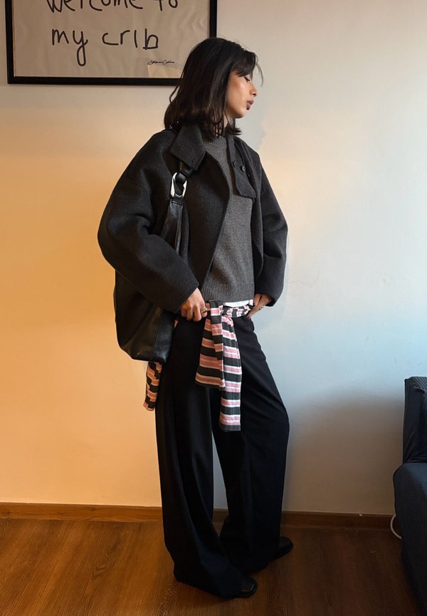 Donna con giacca e maglione grigio scuro, pantaloni larghi neri e camicia a righe legata in vita, in piedi all'interno vicino a una parete con testo incorniciato.