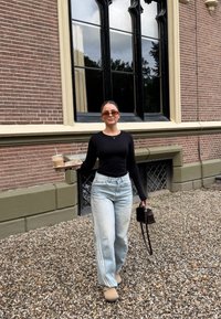 Schwarzes langärmliges Oberteil, hellblaue weite Jeans mit seitlichen Streifen, beige Clogs, hält ein Eis-Kaffee und eine kleine schwarze Tasche, steht auf Kies.