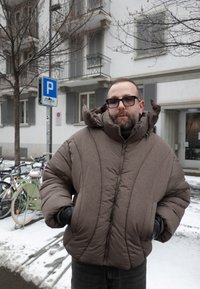 Άνδρας με καφέ μπουφάν και μαύρα γάντια στέκεται σε χιονισμένο πεζοδρόμιο κοντά σε ποδήλατα και πινακίδα στάθμευσης σε αστική κατοικημένη περιοχή.