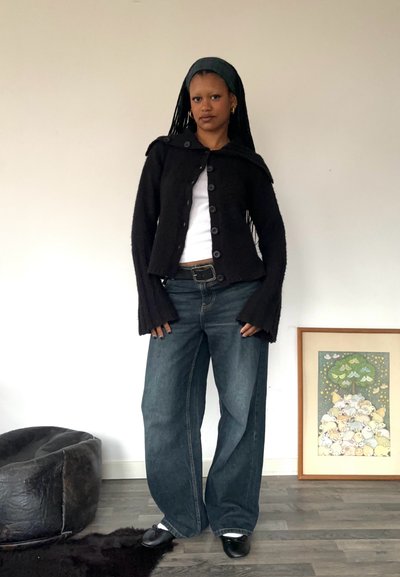 Cardigan negro de lana con botones grandes y mangas anchas, llevado sobre una camiseta blanca. Unos vaqueros azul holgados y zapatos negros completan el atuendo.