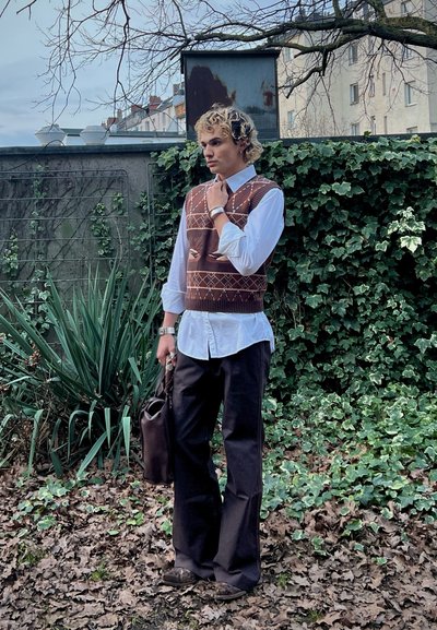 Joven con cabello rizado que lleva un chaleco marrón de patrón, camisa blanca, pantalones negros, sosteniendo una bolsa oscura, de pie al aire libre junto a una pared cubierta de hiedra.