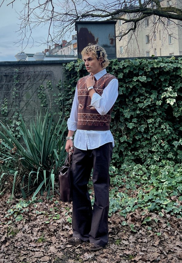 Ung person med krøllete hår iført brun mønstret genservest, hvit skjorte, svarte bukser, holder en mørk veske, står utendørs ved en vegg dekket av eføy.