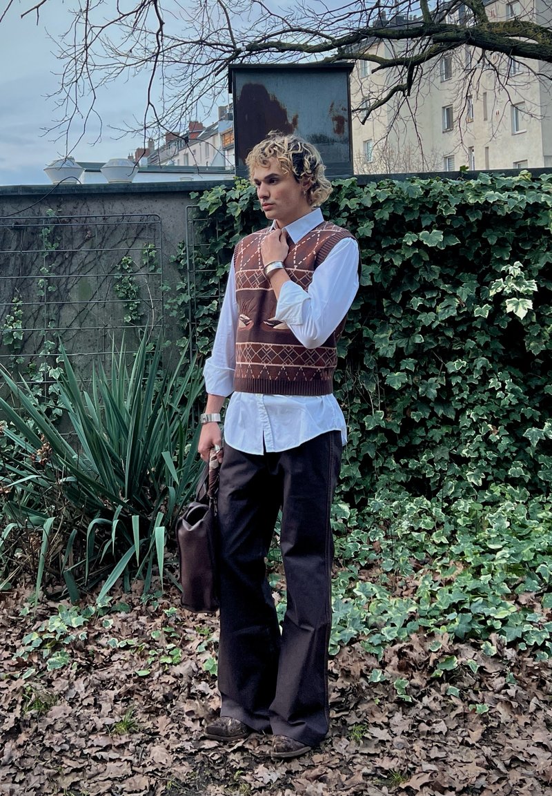 Jeune personne aux cheveux bouclés portant un gilet marron à motifs, une chemise blanche, un pantalon noir, tenant un sac sombre, debout en plein air près d'un mur couvert de lierre.