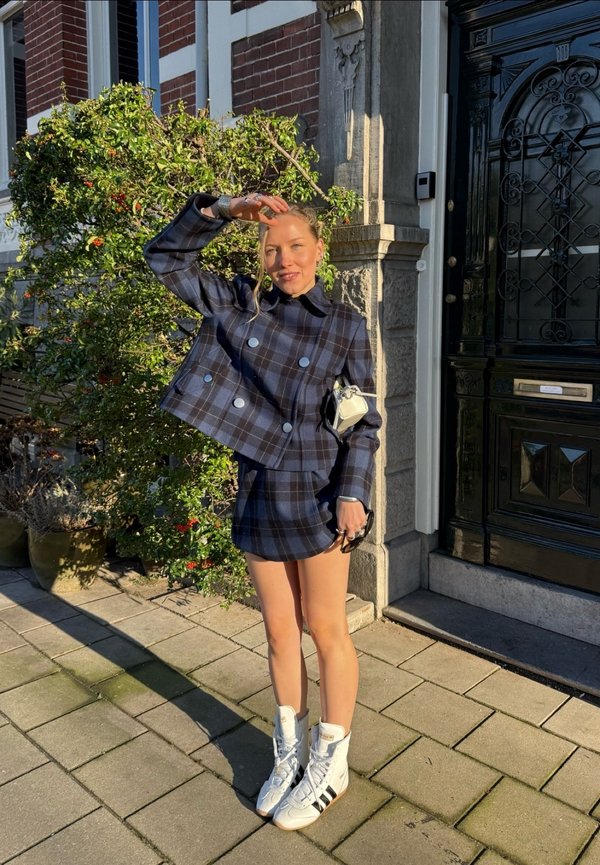 Femme se protégeant les yeux du soleil, portant une veste à carreaux bleus et une jupe, des baskets blanches montantes, debout sur le trottoir près d'une porte noire et de verdure.