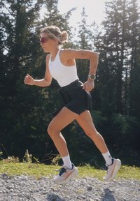 Mulher atlética a correr numa estrada de cascalho, vestindo uma camisola sem mangas branca, calções pretos e sapatilhas de corrida rosas com meias brancas.