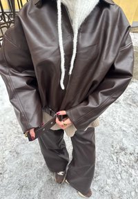 Personne portant une veste en cuir marron foncé surdimensionnée et un pantalon ajustant la ceinture de la veste tout en se tenant sur un sol enneigé en plein air.