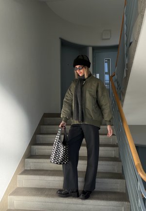 Chaqueta bomber verde oliva, bufanda negra, pantalones negros de talle alto, bolso negro con lunares blancos, zapatos negros, en unas escaleras en un pasillo bien iluminado.