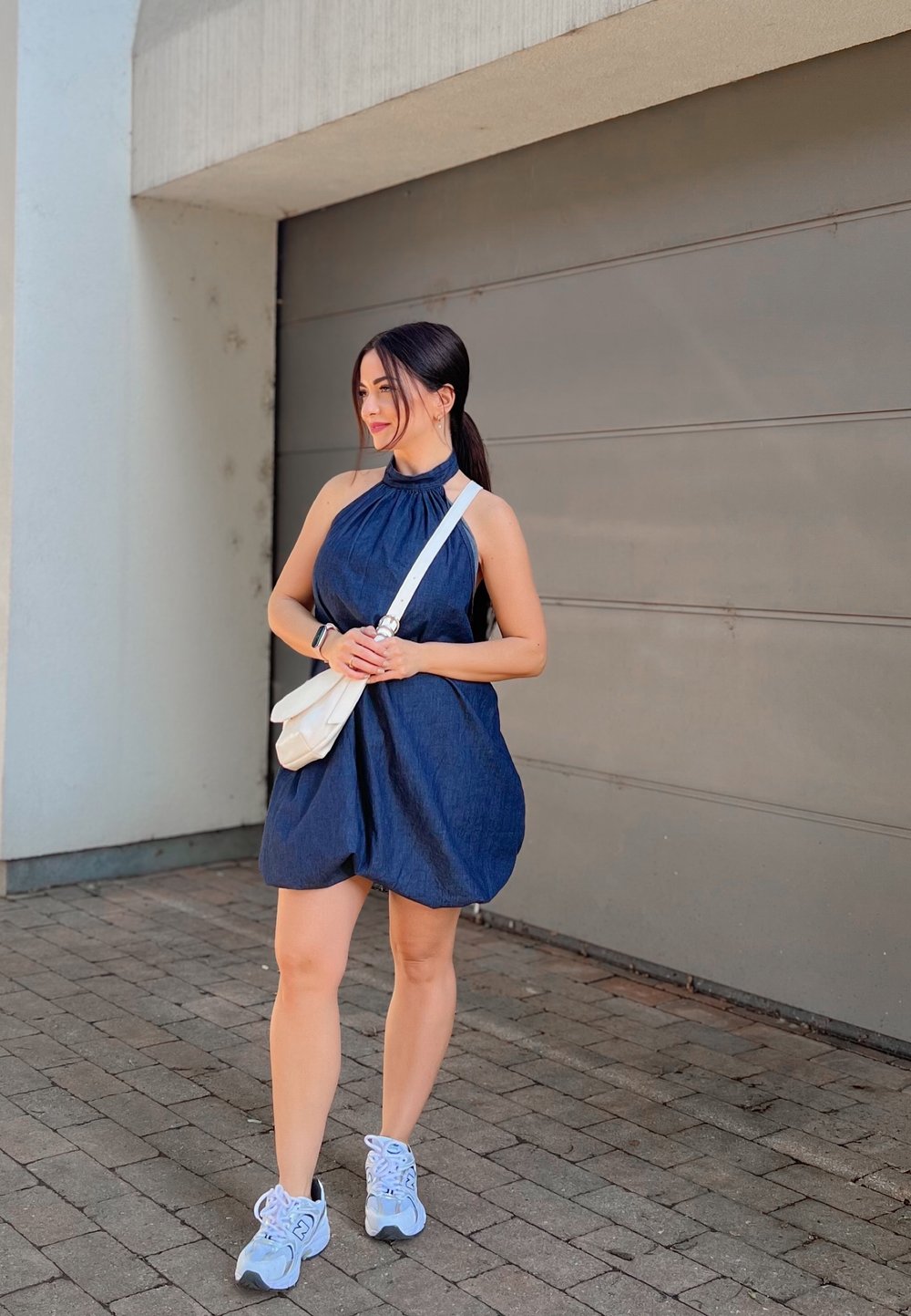A woman wearing a sleeveless blue dress and white sneakers stands on a paved footpath, holding a white crossbody bag, in front of a grey garage door.
