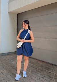 A woman wearing a sleeveless blue dress and white sneakers stands on a paved footpath, holding a white crossbody bag, in front of a grey garage door.