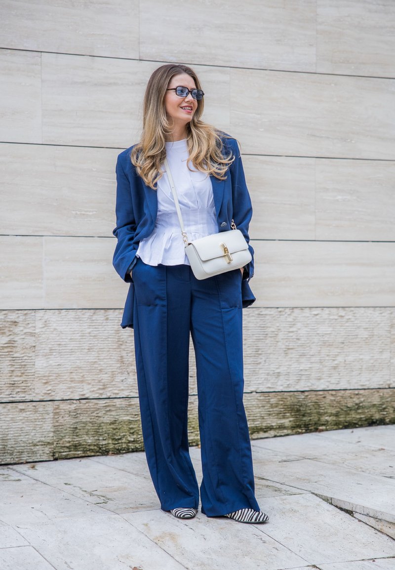 Femme aux longs cheveux portant un costume bleu, un chemisier blanc, des chaussures rayées, des lunettes de soleil et un sac bandoulière blanc, debout contre un mur de pierre.