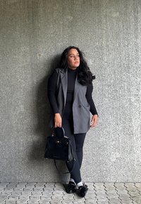 Grå ermeløs vest over en svart turtleneck, svarte leggings, blanke svarte sko, holder en svart håndveske med sølvfester, stående mot en grå vegg.