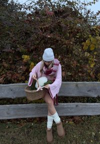 Roze gebreide trui met uitlopende mouwen, witte gebreide muts, geruite sjaal en bruine enkellaarsjes. Houdt een rieten mand met groen en decoratie vast.