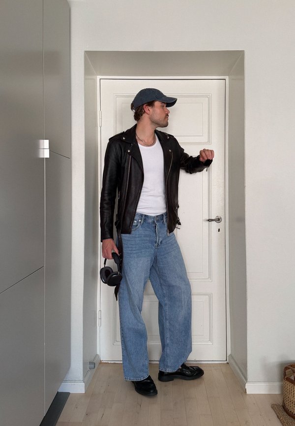 Homme en veste en cuir noire, chemise blanche, jean bleu et casquette, debout à l'intérieur, tenant un casque audio et regardant une porte blanche fermée.