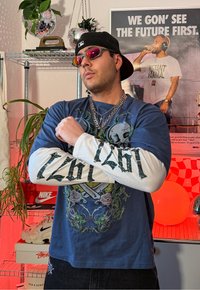 Jeune homme portant des chemises graphiques superposées, des chaînes, des lunettes de soleil et une casquette à l'envers, debout les bras croisés dans une pièce avec des plantes et une affiche motivante.