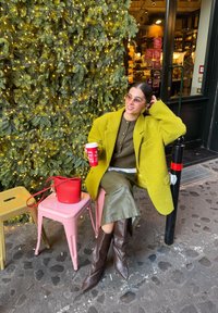 Manteau vert vif texturé, haut vert olive, jupe argent métallique, bottes marron montant jusqu'aux genoux, tenant une tasse rouge. Tabourets métalliques roses et jaunes.