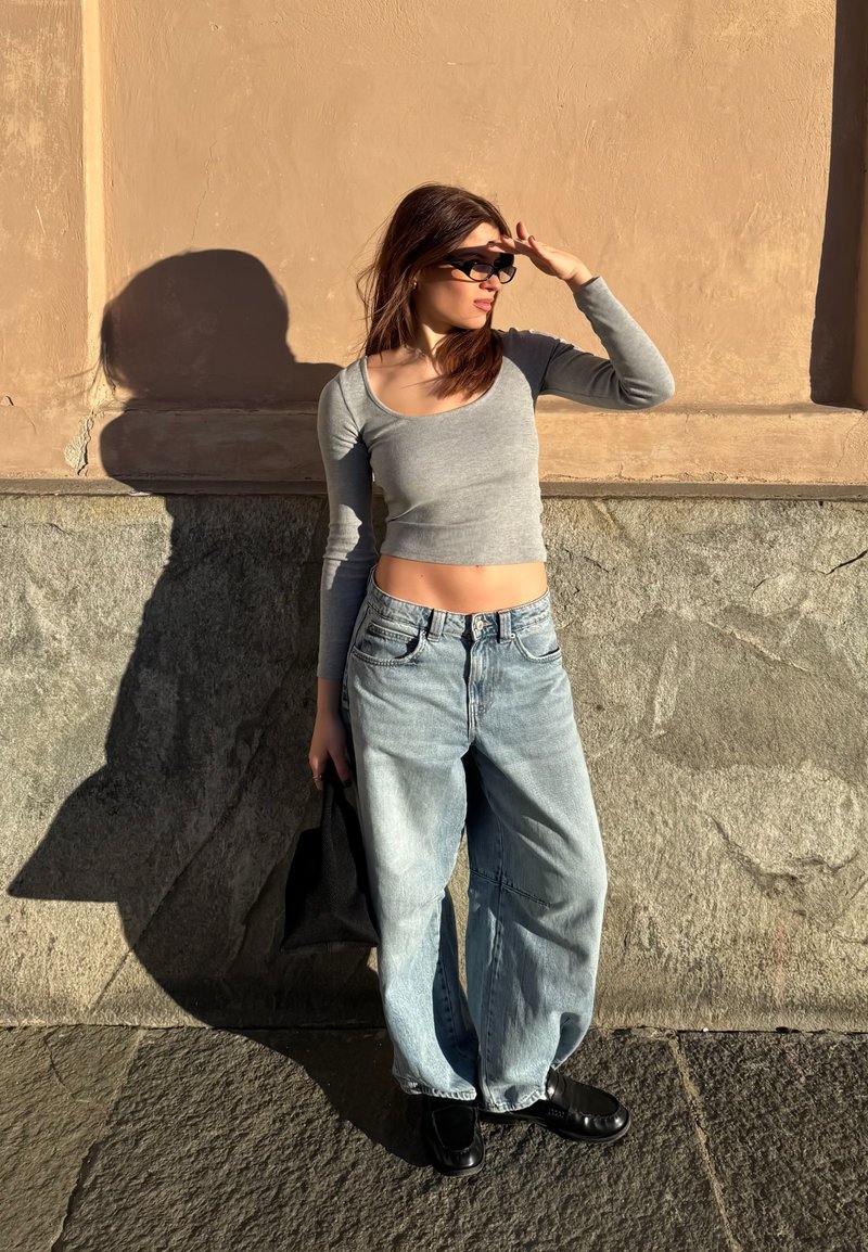 Jeune femme en haut court gris, jean large et chaussures noires, protégeant ses yeux avec des lunettes de soleil contre un mur en pierre éclairé par le soleil.