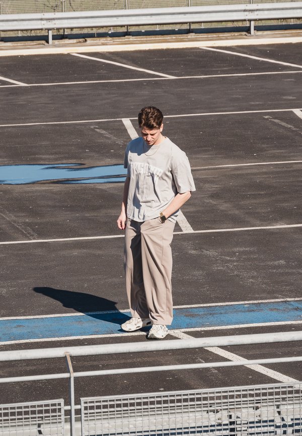 Camiseta de manga corta gris claro con texto, pantalones anchos beige y zapatillas blancas. De pie sobre asfalto con líneas de estacionamiento azules, proyectando una sombra.