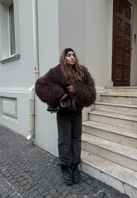 Manteau oversize marron et duveteux, jean foncé, bottes plates-formes noires. Tenant un sac marron, debout sur des marches en pierre près d'un mur beige.