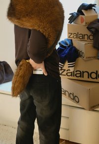 Brun pelsimitation krave draperet over en mørk skjorte, med en matchende pelsimitation hale. Sorte bukser med et synligt mærke. Zalando kasser i baggrunden.