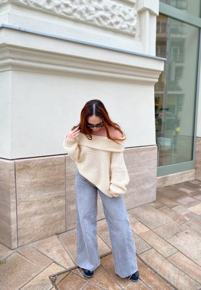 Jersey de punto oversize color crema con diseño de hombros al descubiertos, combinado con jeans gris de pierna ancha y calzado negro, sobre un pavimento de piedra.