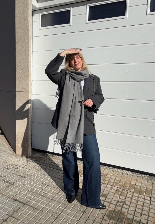 Vrouw staat op betegeld trottoir en beschut haar ogen tegen de zon, draagt een grijze sjaal, een donker colbert, wijde jeans en zwarte puntige schoenen bij een witte garagedeur.