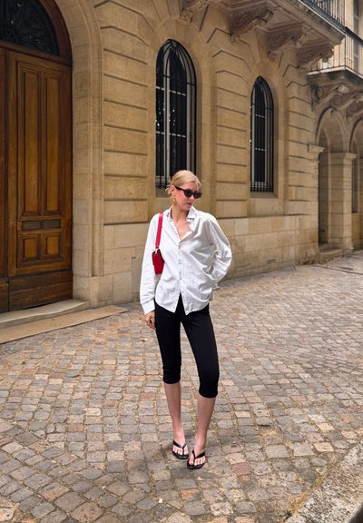 Mujer rubia con gafas de sol, camisa blanca, pantalones capri negros y chanclas, de pie en una calle empedrada junto a un edificio de piedra con ventanas arqueadas.