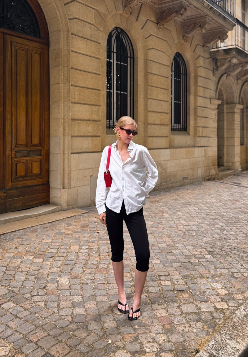 Mujer rubia con gafas de sol, camisa blanca, pantalones capri negros y chanclas, de pie en una calle empedrada junto a un edificio de piedra con ventanas arqueadas.