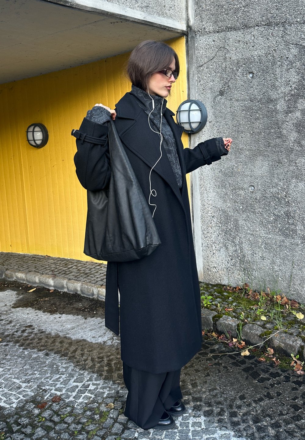 Black wool coat with wide lapels, teamed with dark trousers. A black leather bag is slung over one shoulder. The background showcases a yellow wall.