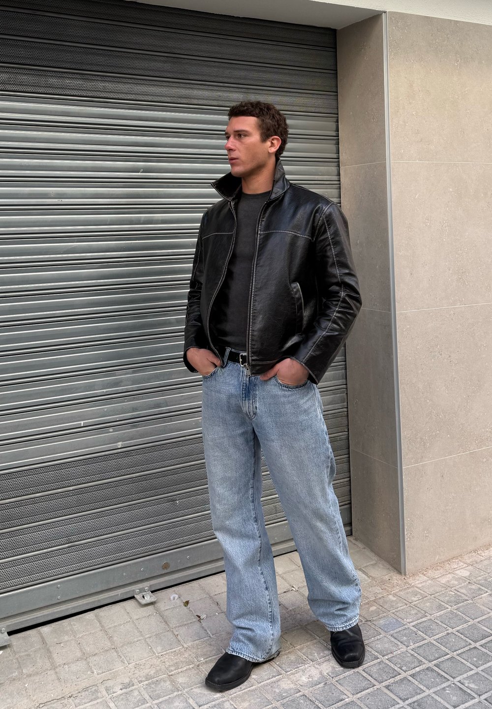 Jeune homme debout les mains dans les poches, portant une veste en cuir noire, une chemise noire, un jean bleu clair et des chaussures noires près d'une grille métallique.