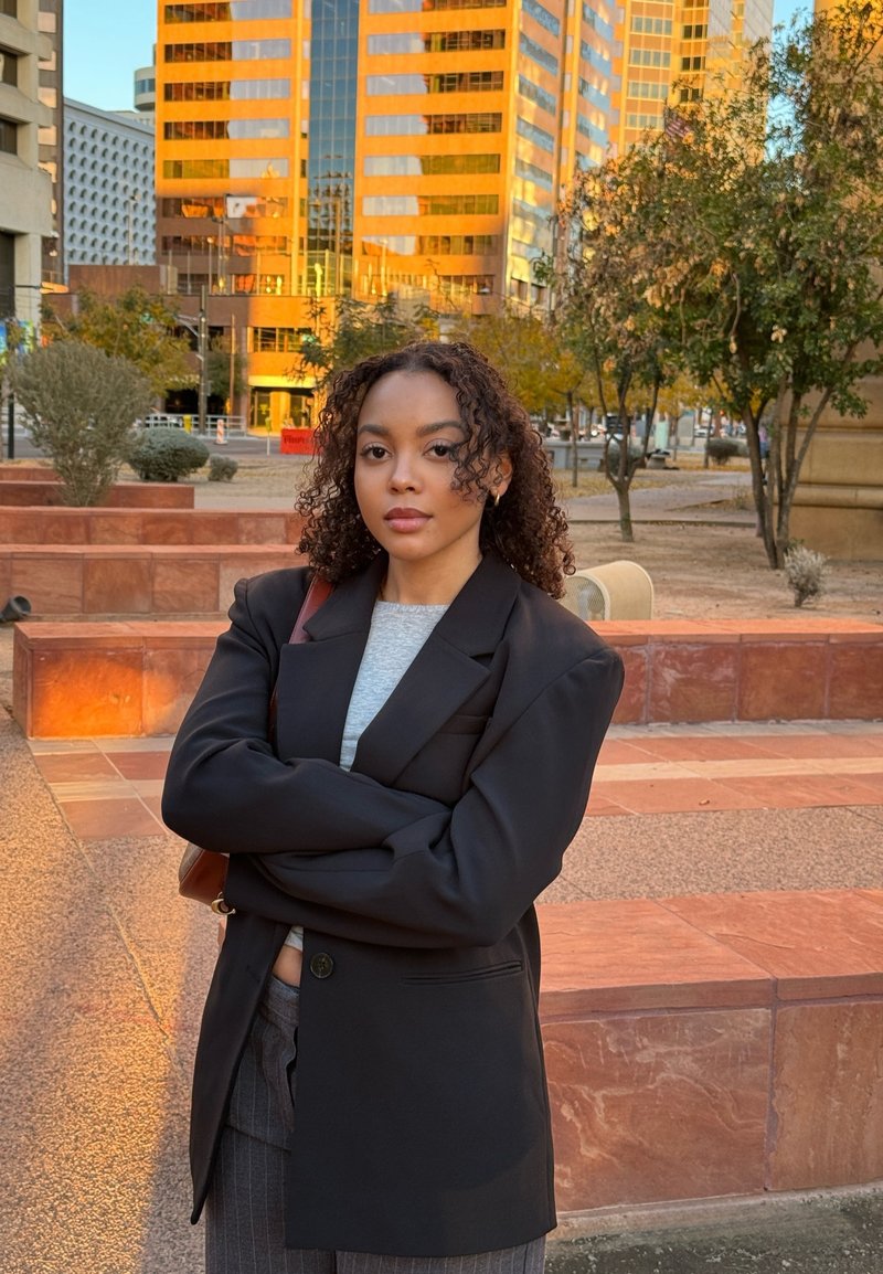 Blazer noir surdimensionné sur une chemise grise, avec un pantalon à rayures. Cheveux bouclés, bras croisés, debout à l'extérieur devant un bâtiment réfléchissant.