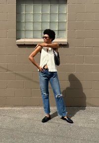 Λευκό ριγέ αμάνικο μπλουζάκι, μπλε τζιν ψηλής μέσης με σκισμένο γόνατο, μαύρες λοφάδες και γυαλιά ηλίου, πόζαρε μπροστά σε έναν γκρι τοίχο από τούβλα.